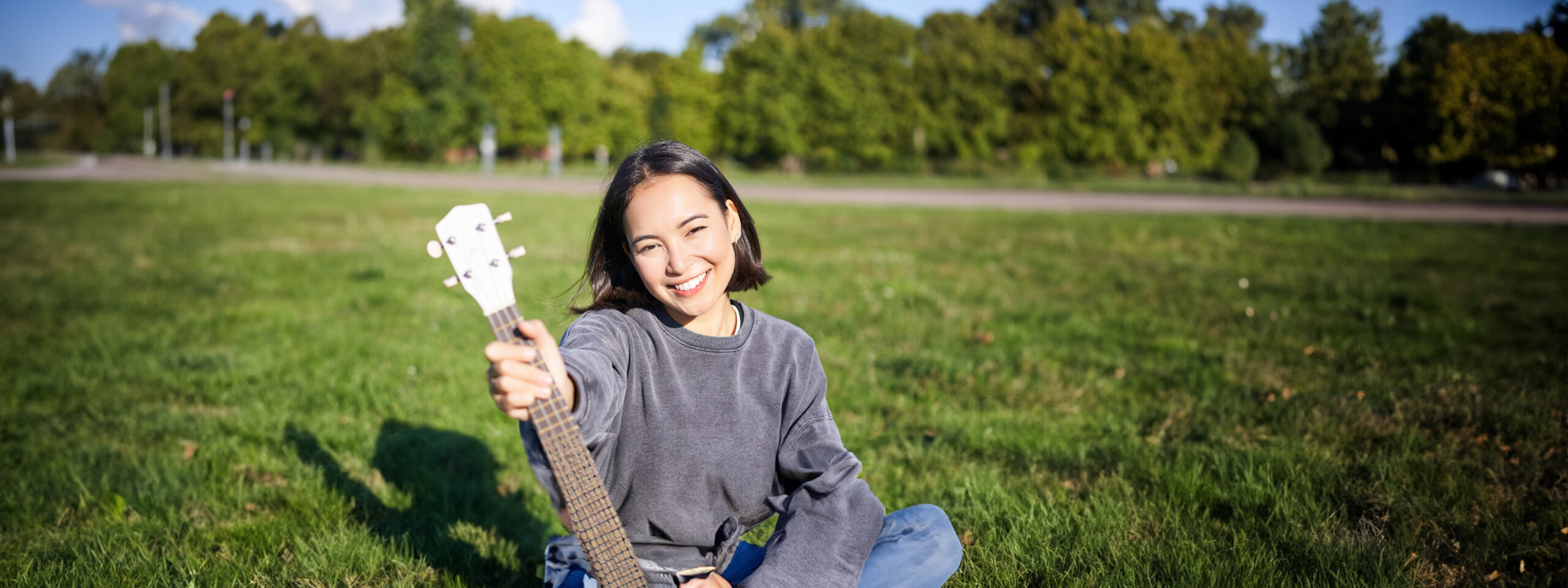 music and instruments. smiling asian girl shows her white ukulele, sits in park and plays small guitar